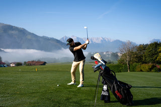 Golfer teeing off on green golf course with mountains in background and golf bag in foreground