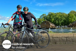 Two cyclists in gear posing with bikes outdoors near a pond and fountain, bright sunny day.