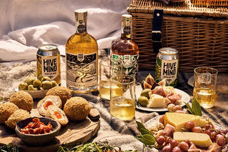 Picnic with Hive Mind mead bottles, cans, glasses, cheese, grapes, and snacks on blanket outdoors