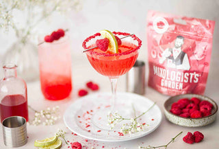 Raspberry margarita cocktail in a glass with red sugar rim, lime, and raspberries, surrounded by fresh raspberries and drink mixing ingredients.